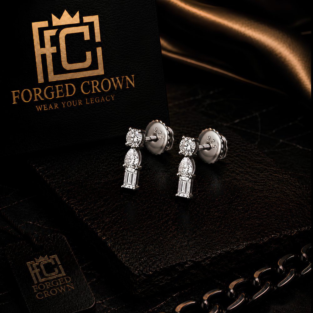 Moissanite 3 Stone Mixed Cut drop stud earrings in platinum plated sterling silver with round, pear and baguette stones in vertical arrangement
 on a black card with 'Forged Crown' branding.